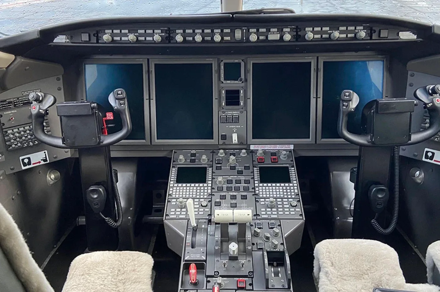The cockpit of a business jet, featuring multiple large display screens, control yokes, and a central control pedestal.