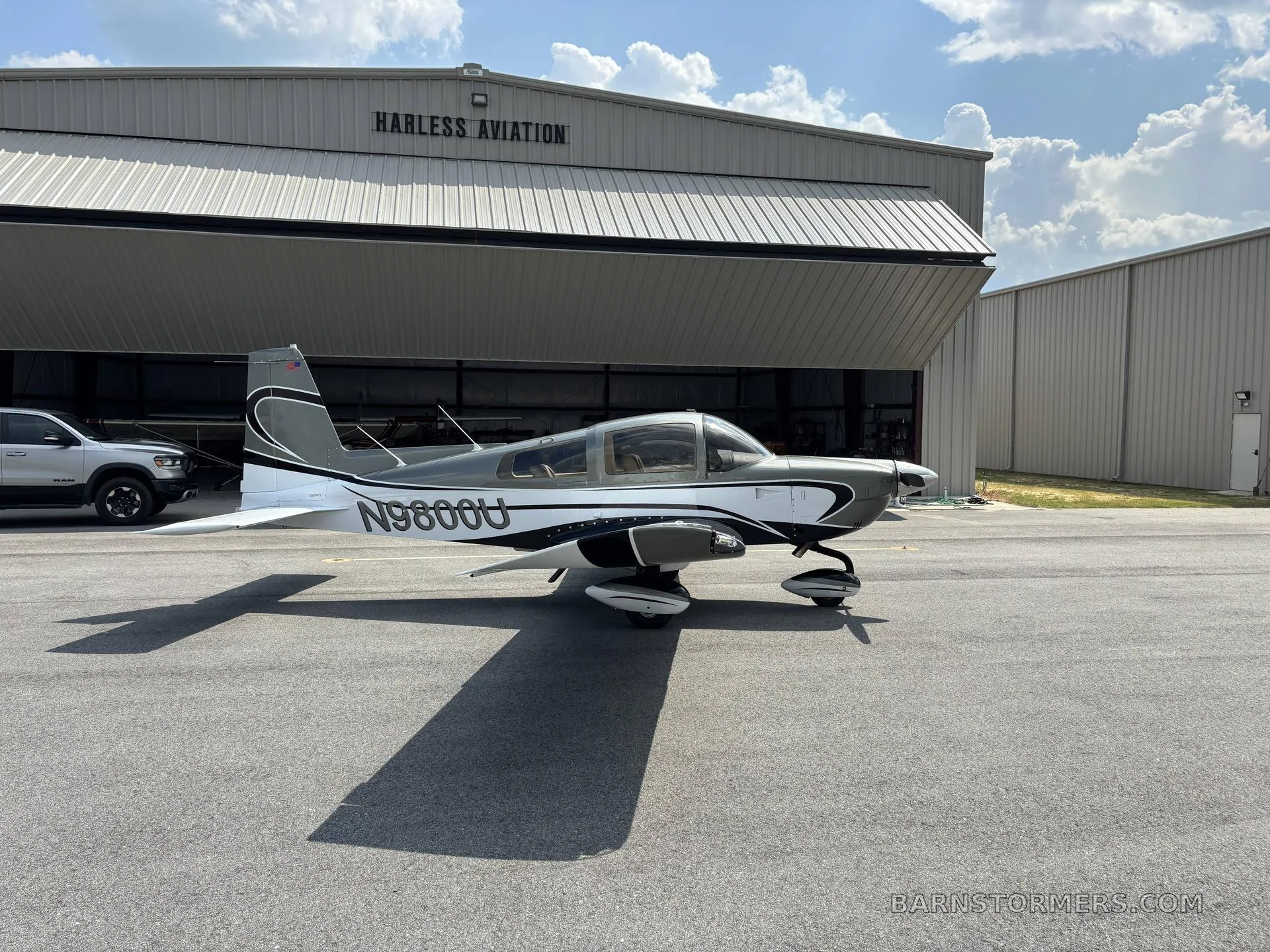 A gray and white Grumman American AA-5B Tiger (N9800U) parked on an airfield in front of a large hangar.