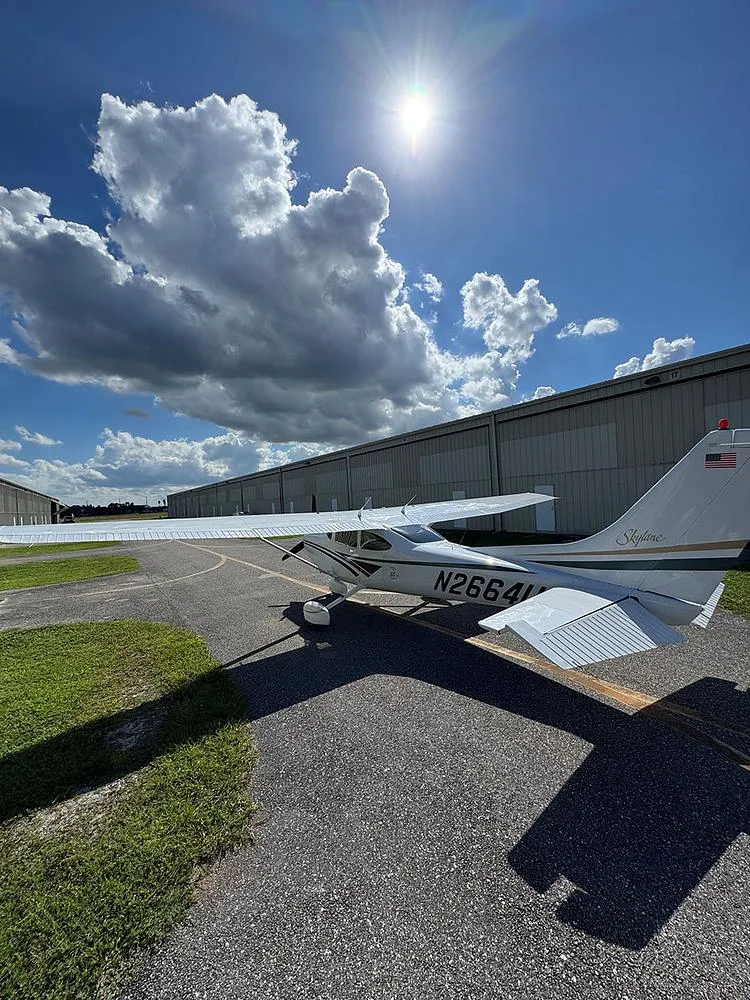 Rear-left view of a white Cessna 182 Skylane with 'N2664U' on the tail, parked on an asphalt ramp with hangars in the background.
