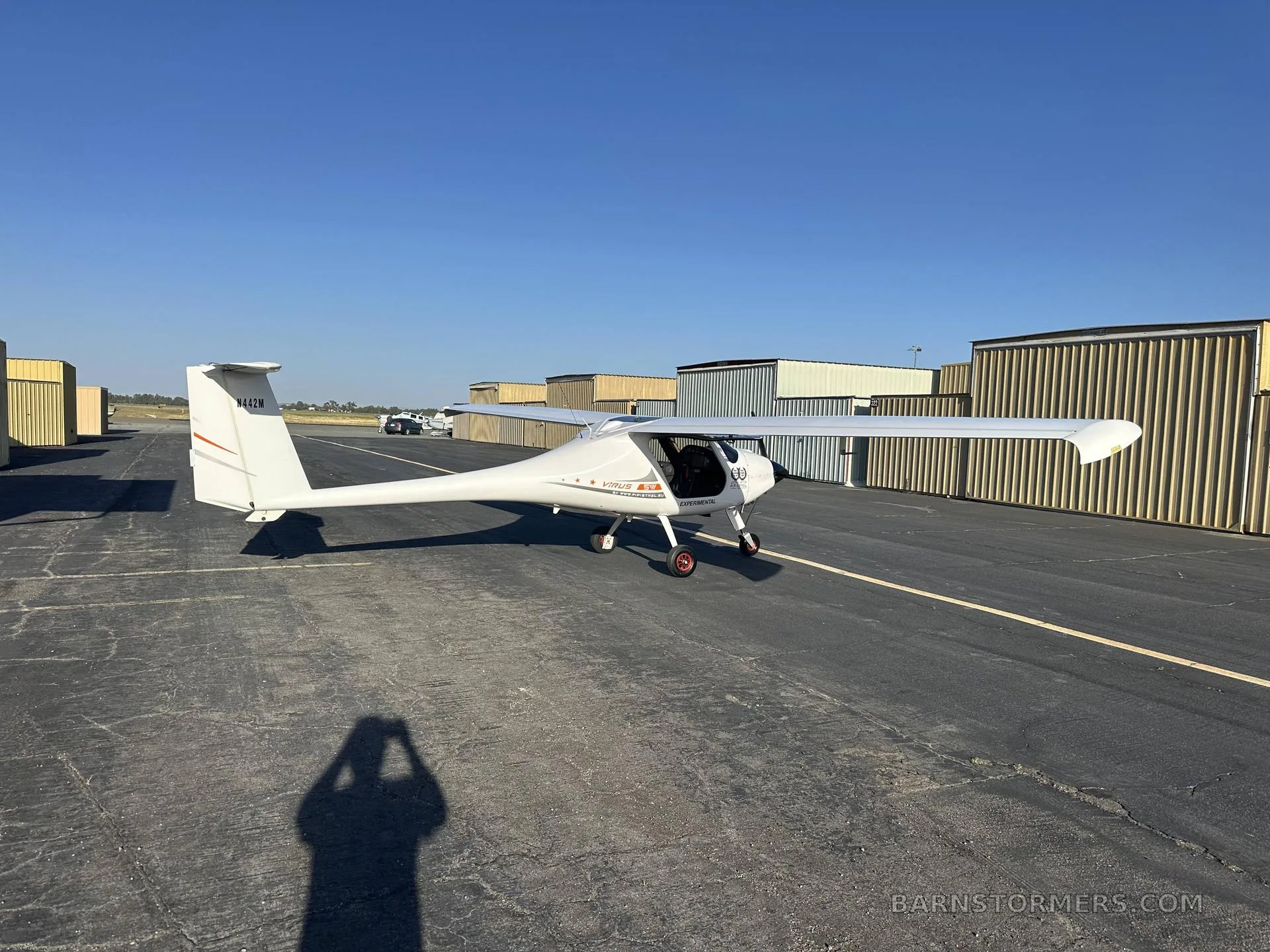 A white light sport aircraft, N442M, with orange stripes on the tail, viewed from the front-right on an asphalt tarmac with hangars in the background.