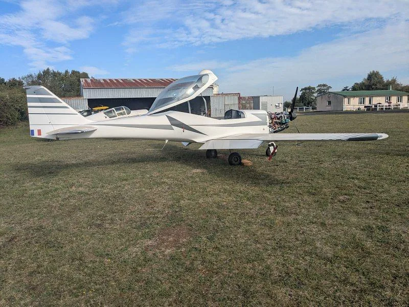 A white light aircraft with its canopy and engine cowling open, parked on a grassy field near a hangar.