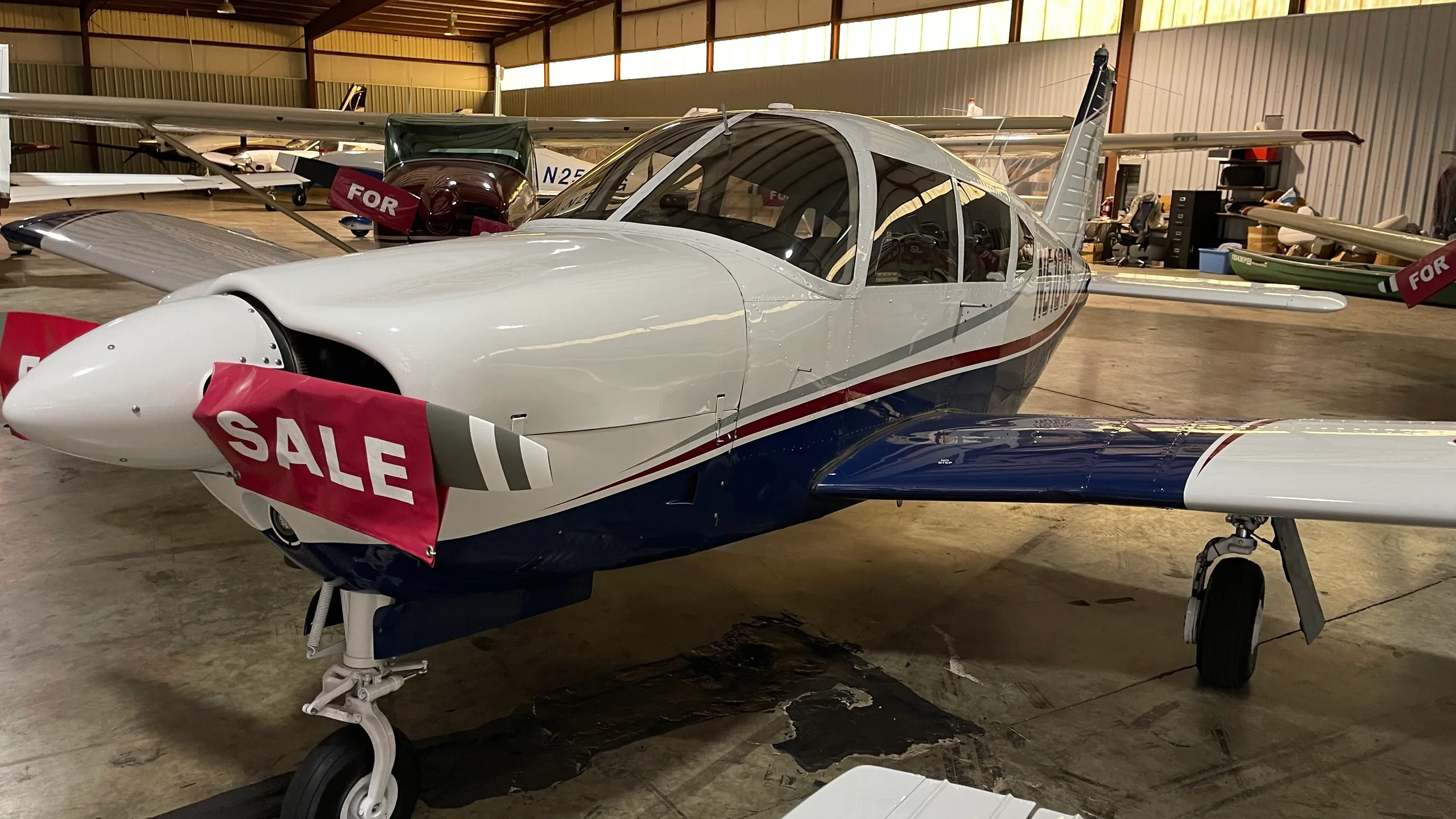 A front-side view of the white and blue Piper PA-28-180 Cherokee Challenger (N5101S) with a red "SALE" banner on its propeller, parked in a hangar.