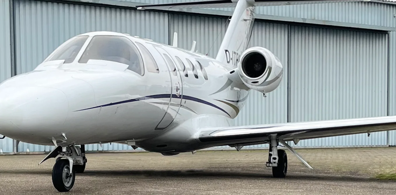 A white Cessna Citation business jet with blue and grey stripes is parked on an apron in front of a hangar.