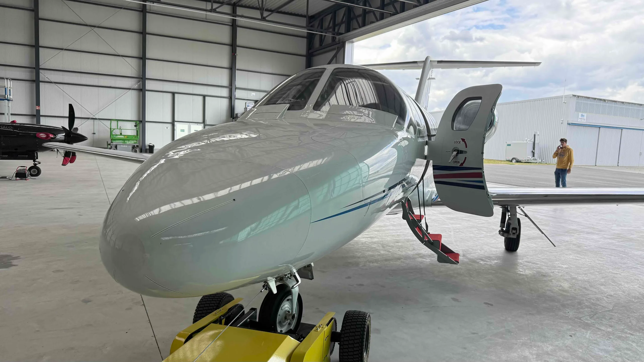 A white private jet, likely a HondaJet, with its main door open and red steps extended, parked inside a hangar with another aircraft partially visible.