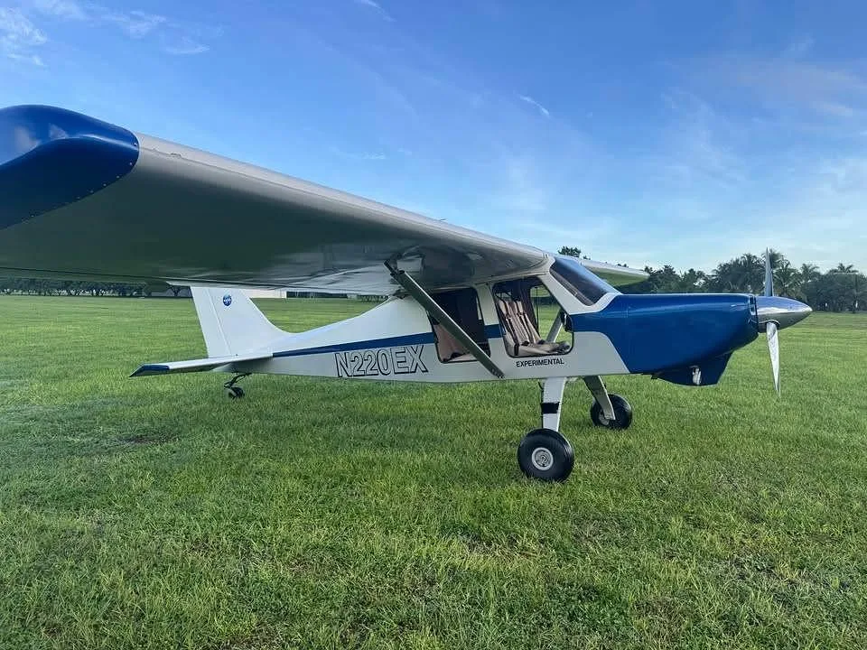 A white and blue experimental aircraft N220EX parked on a green grassy field under a blue sky.