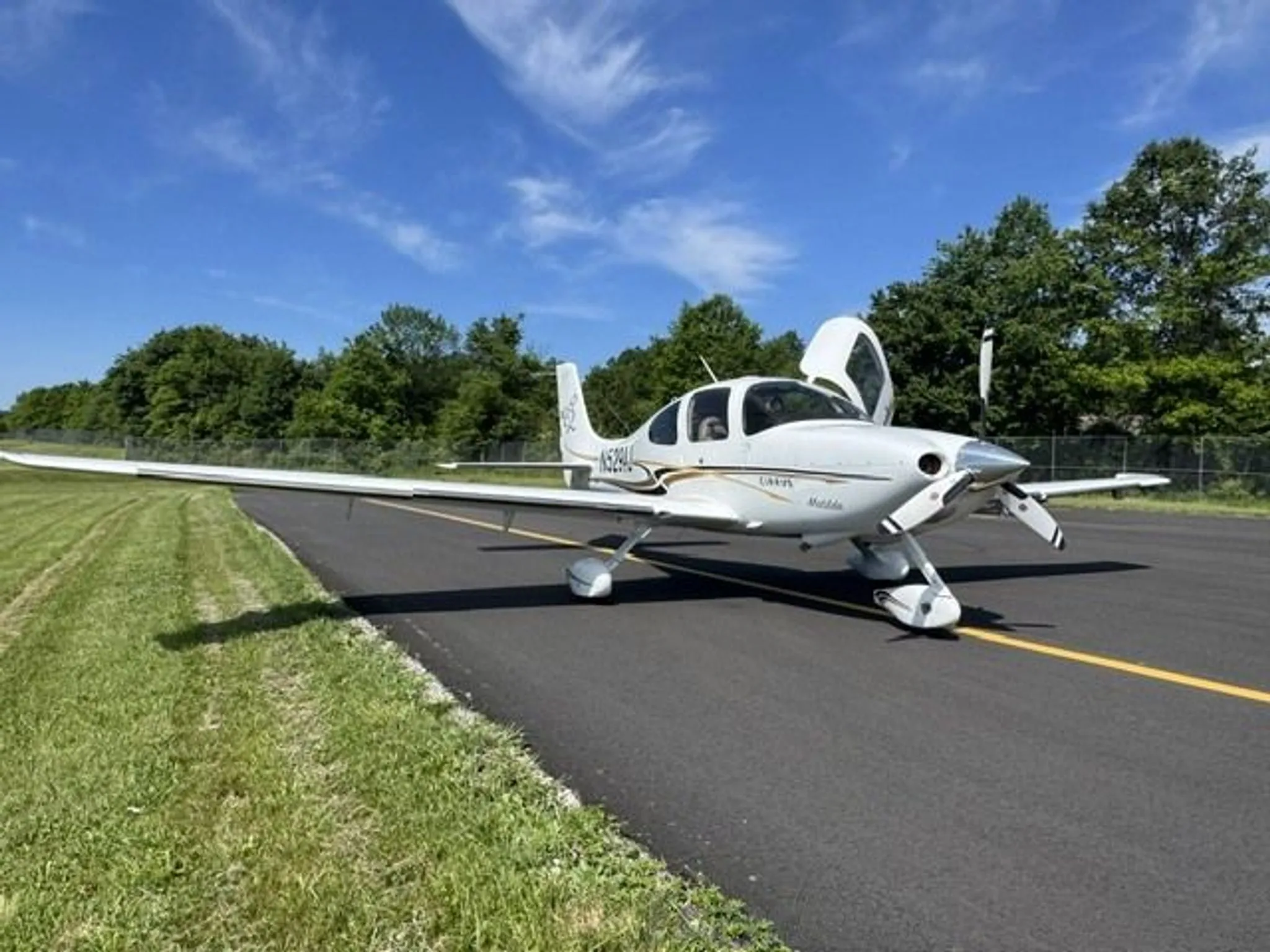 A white Cirrus SR22 aircraft with registration N529AJ and an open door, parked on an asphalt surface next to grass and trees.