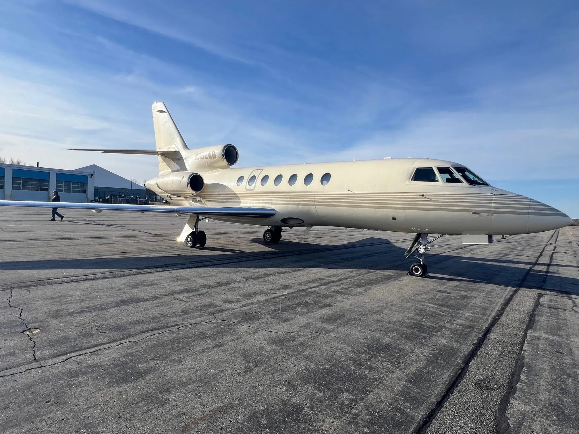 A beige Dassault Falcon business jet with a gold stripe is parked on a tarmac under a blue sky.