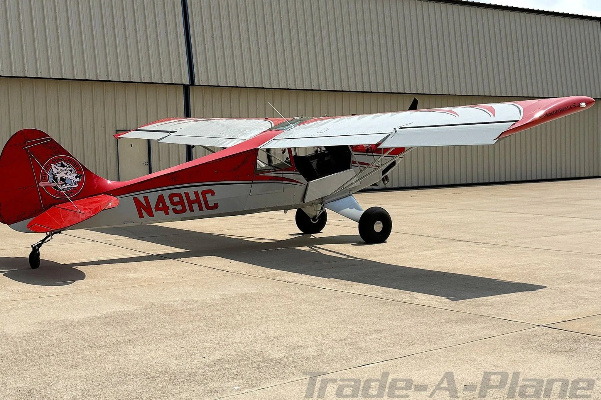 A rear-side view of the red and white Aviat Husky A-1C aircraft, registration N49HC, parked on an airfield.
