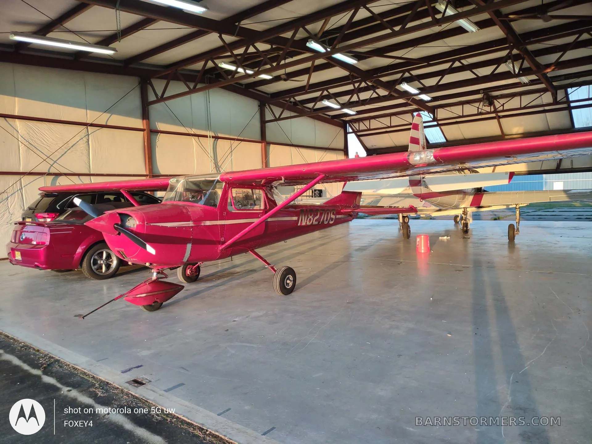 A red Cessna 150 or similar light aircraft with registration N8270S is parked inside a hangar next to a red car.