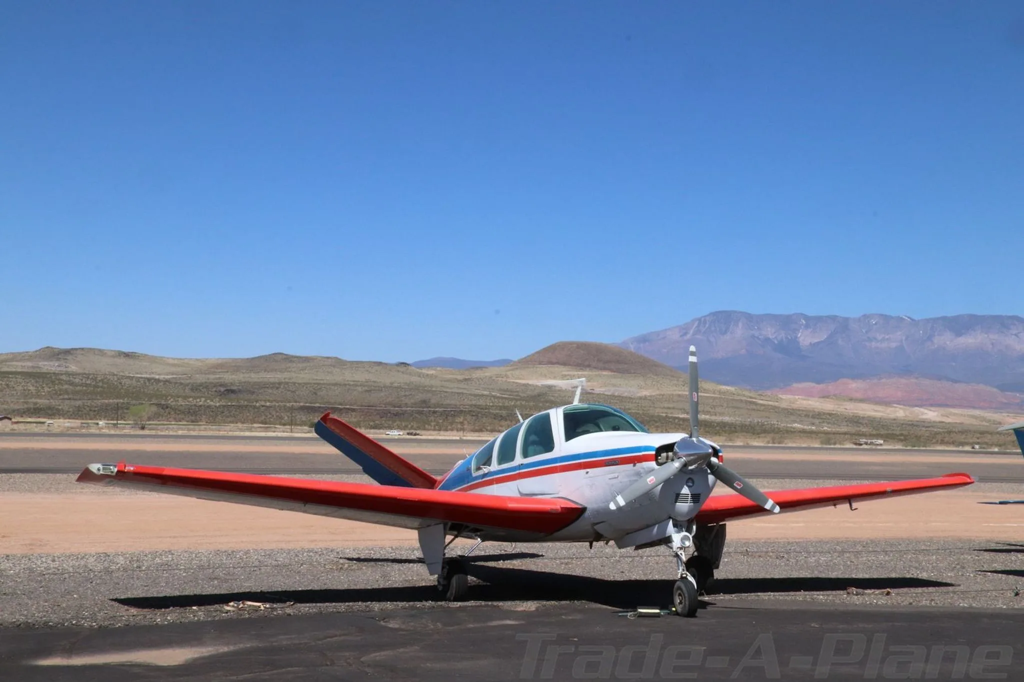 A white, red, and blue Beechcraft Bonanza N7208R is parked on a tarmac, viewed from the front with mountains in the background.