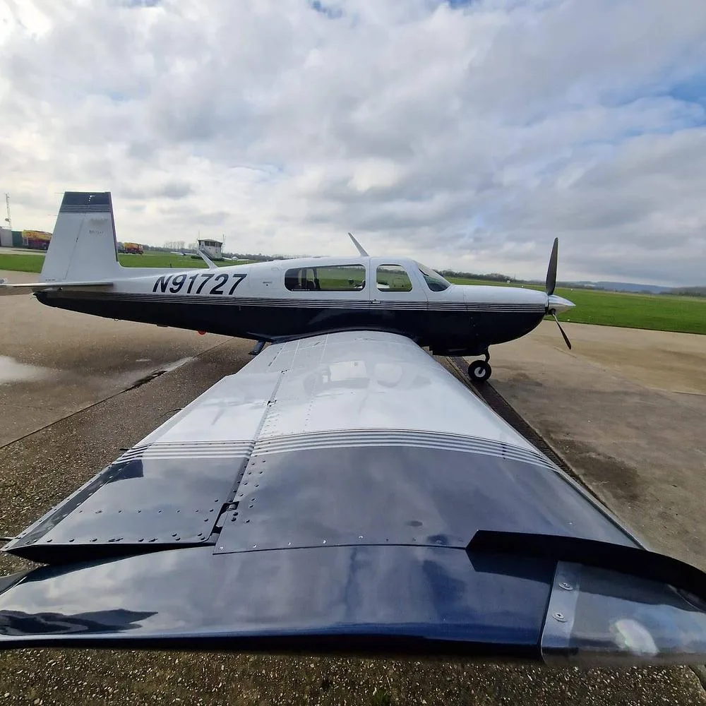 A view from over the wing of a white and blue single-engine aircraft, N91727, parked on an airfield under a cloudy sky.