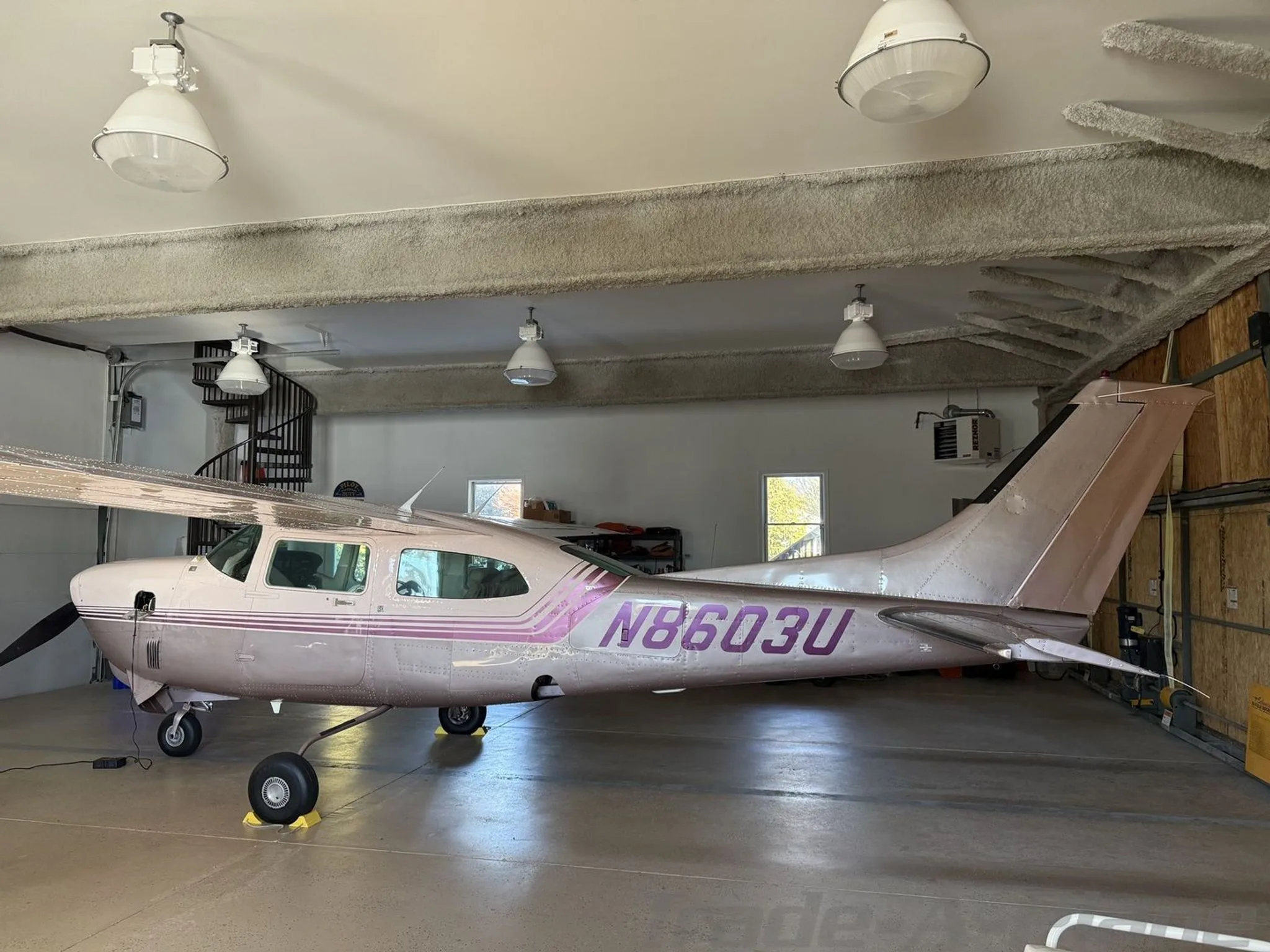 A side view of a light pink and purple Cessna 210 Centurion aircraft, registration N8603U, parked in a hangar.