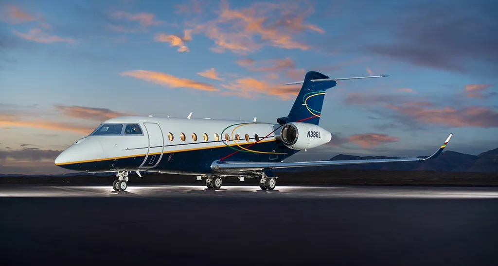 A Gulfstream G550 aircraft, registration N38GL, with a white and dark blue paint scheme and colorful stripes, sits on a tarmac at sunset.