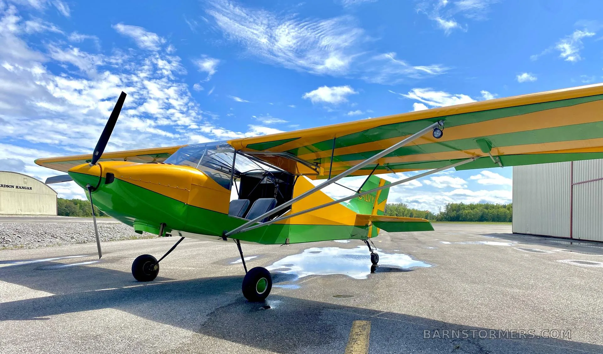 A yellow and green high-wing aircraft with its door open, parked on an asphalt ramp under a blue sky with clouds.