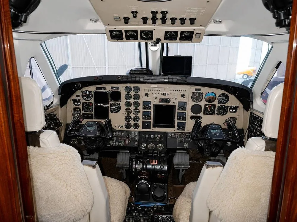 The cockpit features a complex instrument panel with analog gauges, screens, and controls, with two seats covered in white sheepskin.