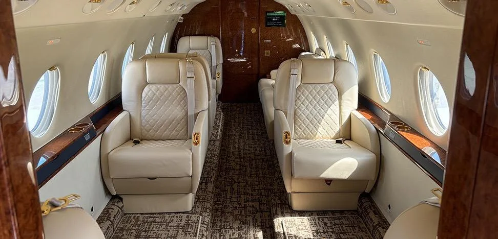 The luxurious interior cabin of a private jet with quilted beige leather seats and dark wood accents.
