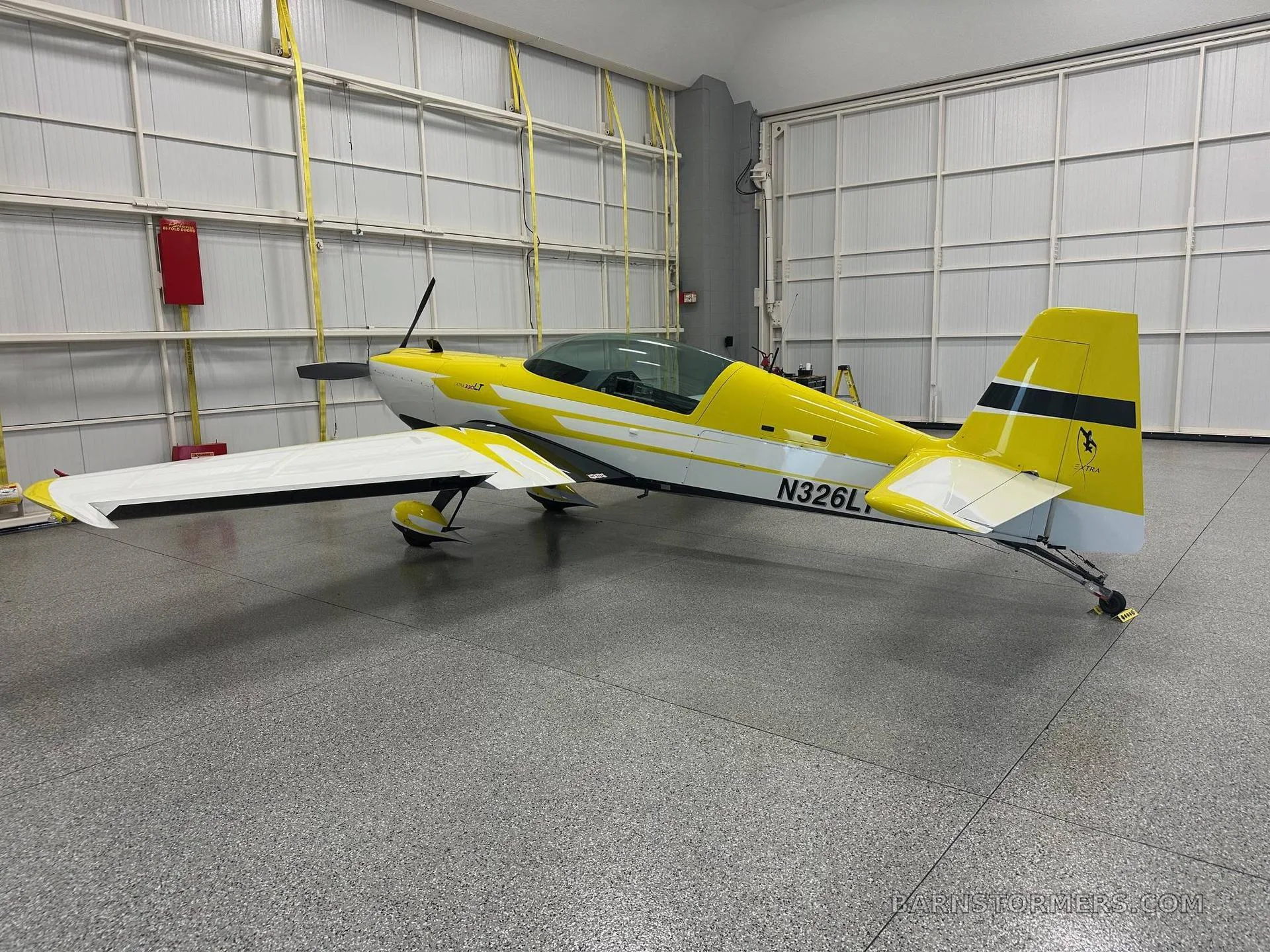 A yellow and white Extra 330LT aerobatic aircraft with registration N326LT, viewed from the rear-left in a hangar.