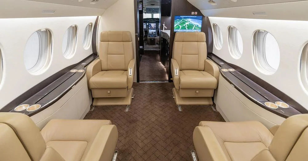 The interior cabin of a business jet features tan leather seats and a screen displaying a map, with a view towards the cockpit.