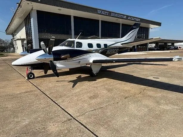 A white and black twin-engine propeller aircraft (N20CG) parked on an airfield in front of the West Houston Airport building.