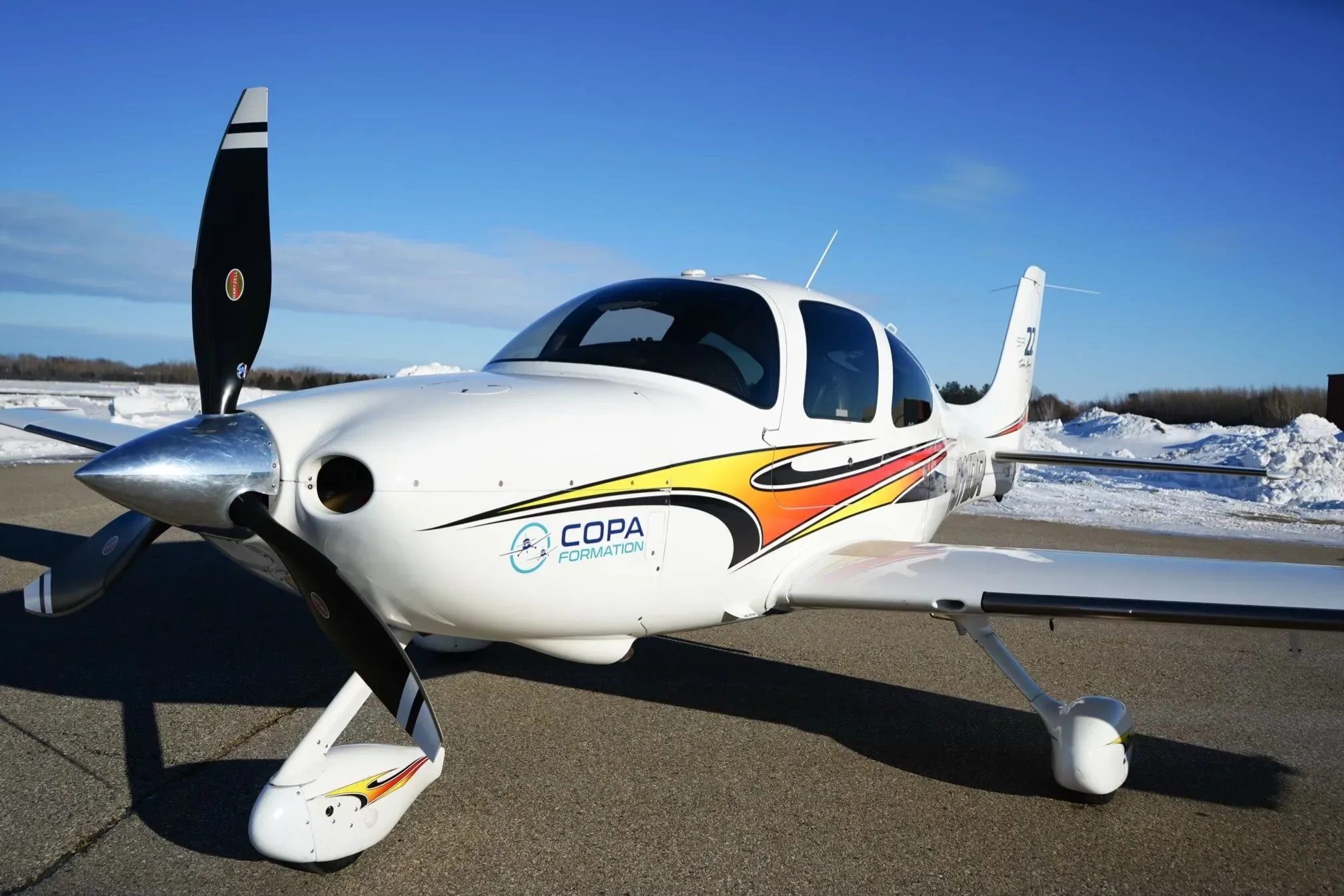 A front-right view of a white Cirrus SR22 aircraft with orange and red stripes and a 'COPA FORMATION' logo, parked on a snowy airfield.