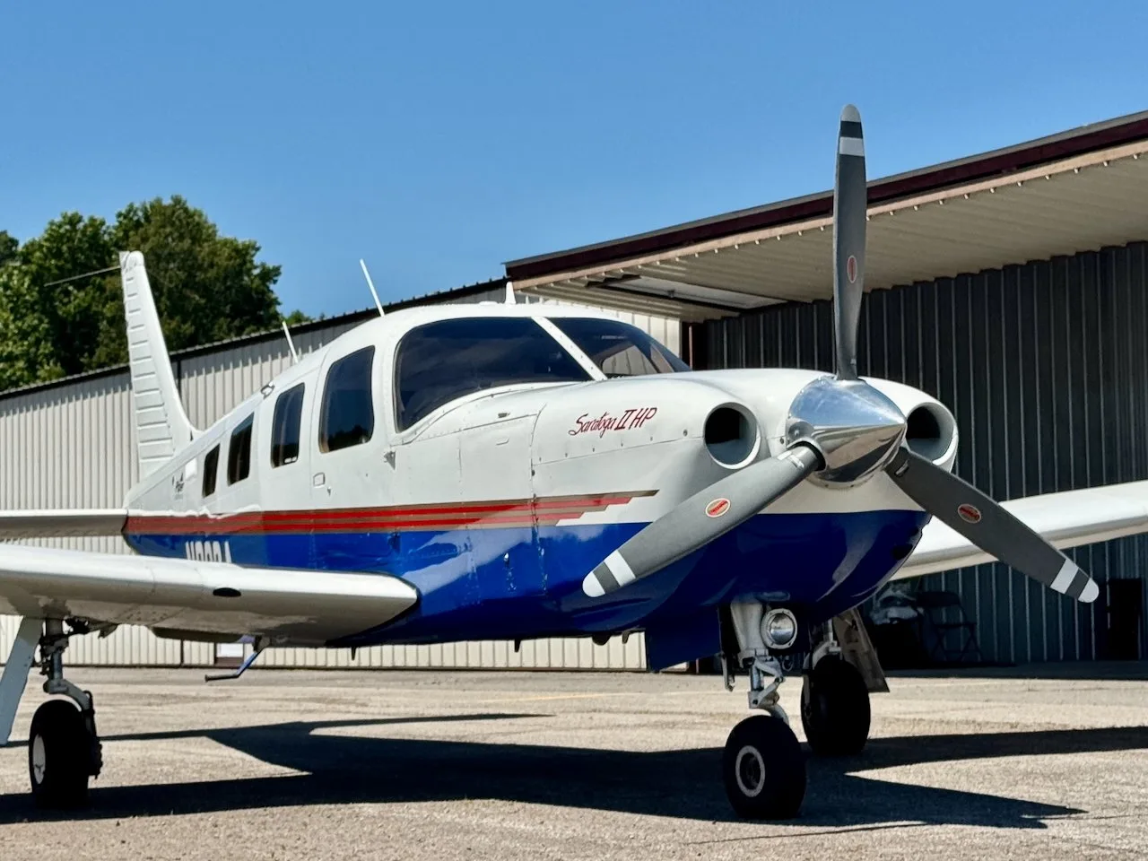A white and blue Piper Saratoga II HP aircraft with red stripes, registration N239A, parked on an airfield with a hangar in the background.