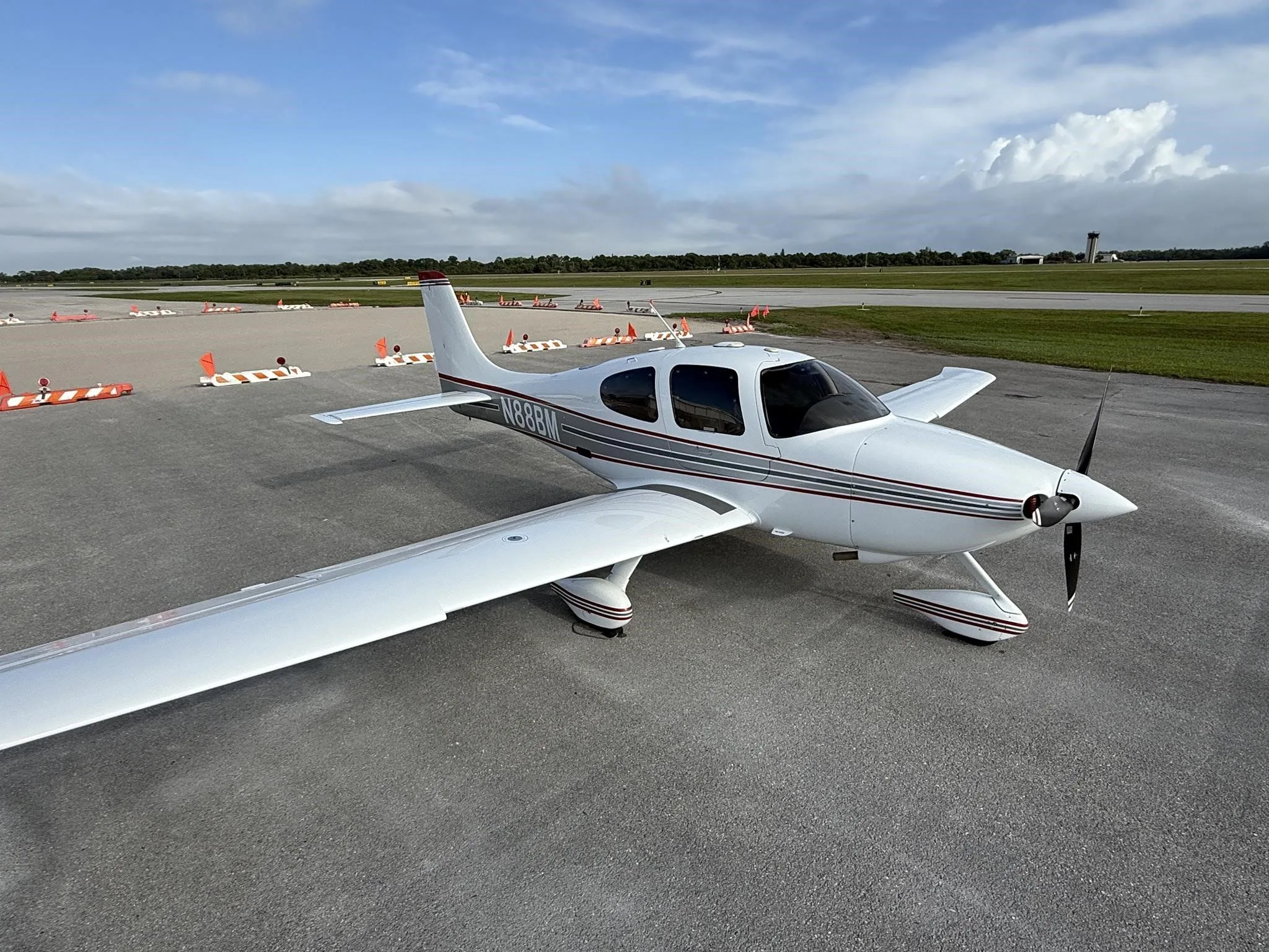 A white Cirrus SR22 aircraft with red and gray stripes, tail number N88BM, parked on an airport tarmac under a partly cloudy sky.