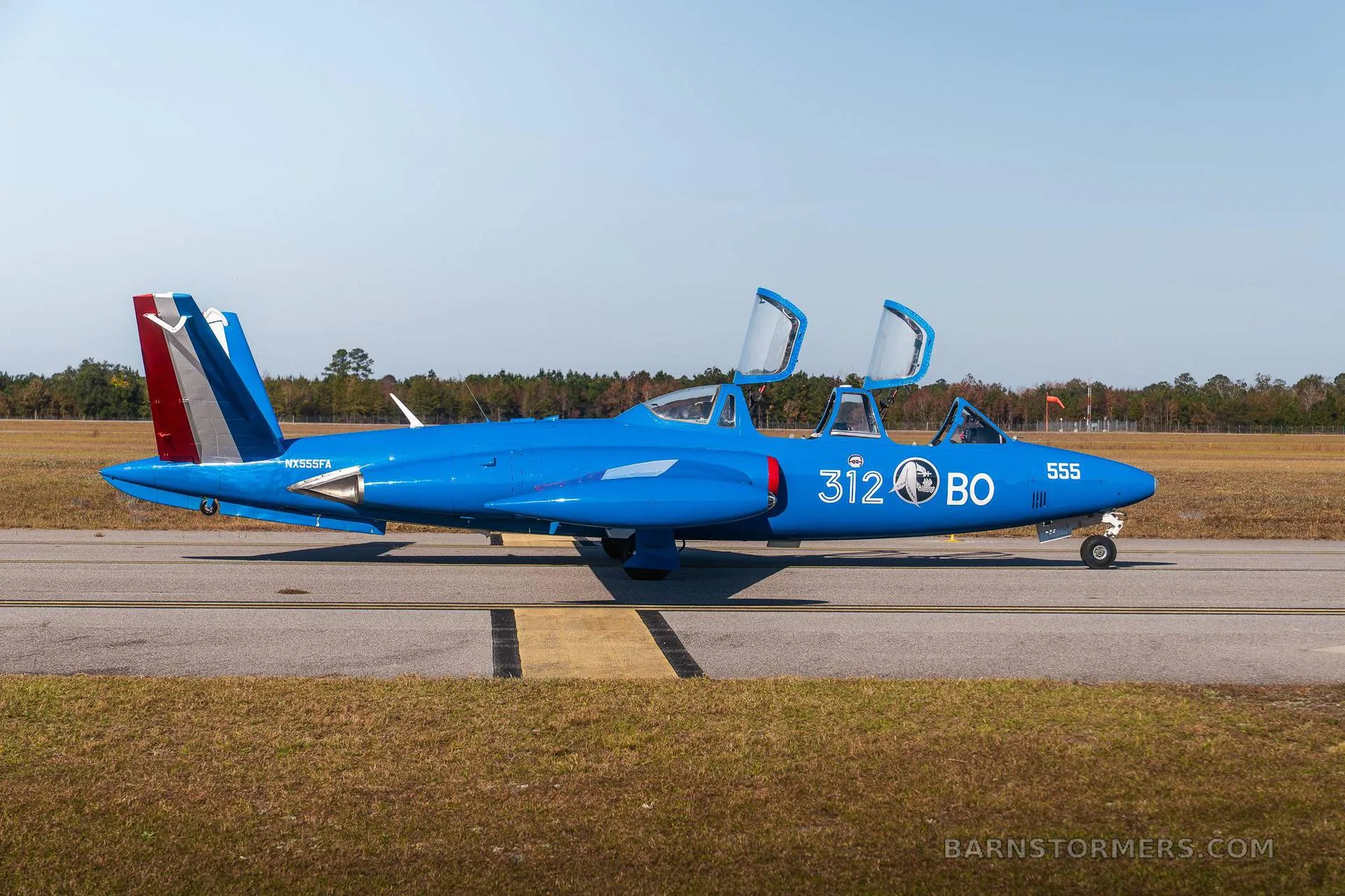 A blue Fouga CM.170 Magister jet aircraft with open canopies is shown from the side on an asphalt runway.