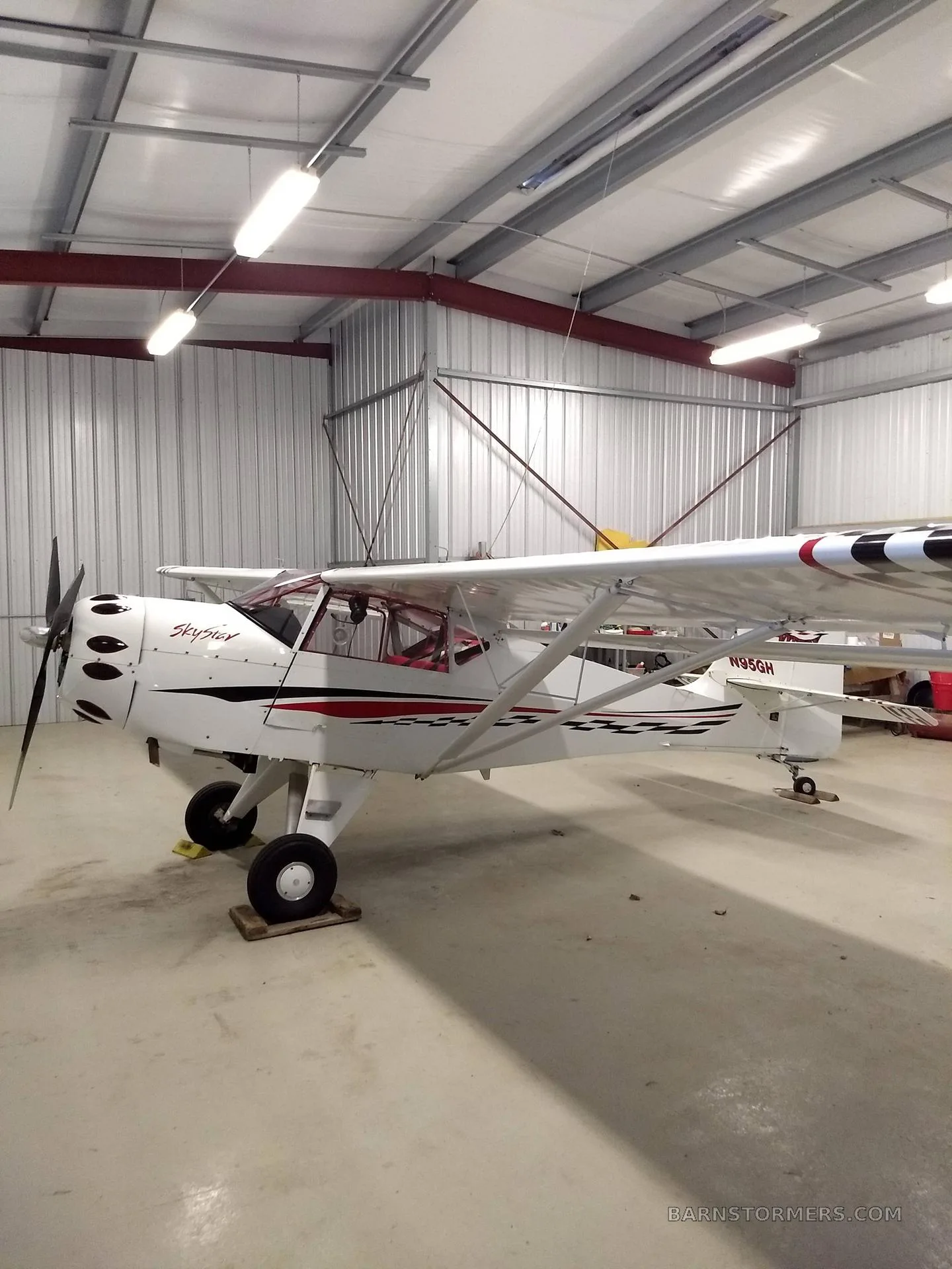 A white SkyStar Kitfox aircraft with red and black checkerboard accents is parked inside a hangar.