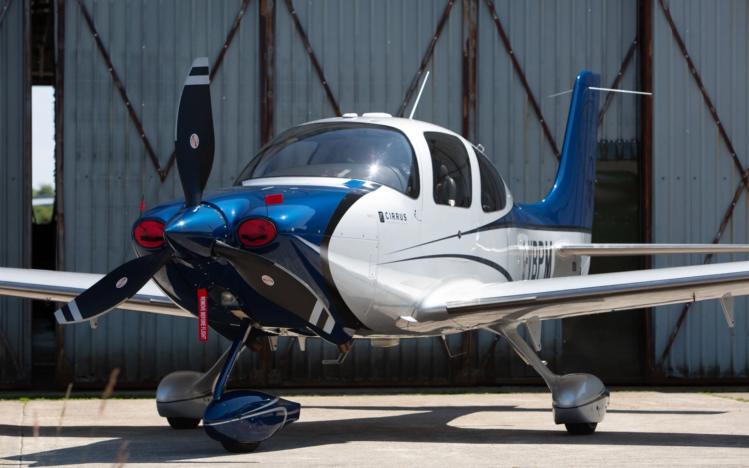 A blue and white Cirrus SR22 aircraft with a propeller, parked in front of a hangar.