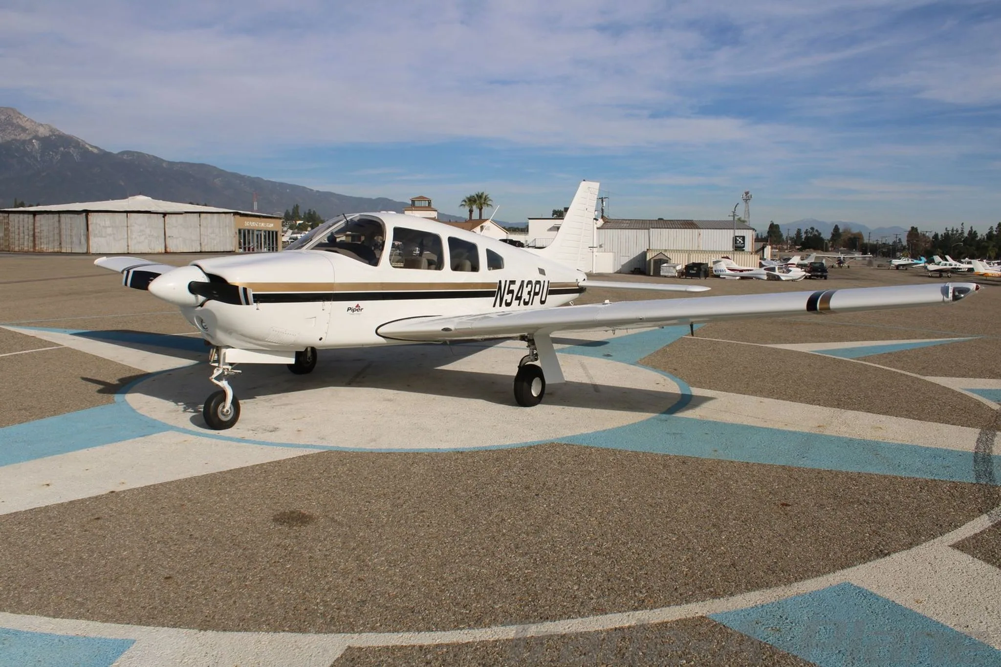 A white Piper aircraft with black and gold stripes, registration N543PU, parked on an airfield with mountains in the background.
