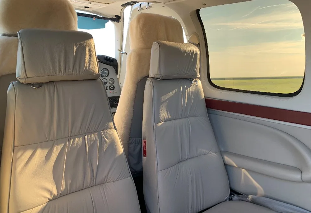 The aircraft cabin features light gray leather seats with a view of the cockpit and exterior through the windows.