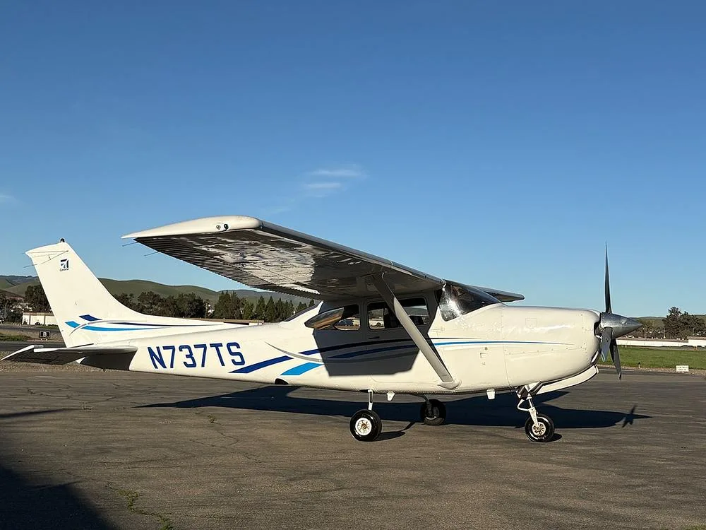 A side view of the white Cessna 182 aircraft with blue stripes, registration N737TS, parked on an airfield under a clear blue sky.