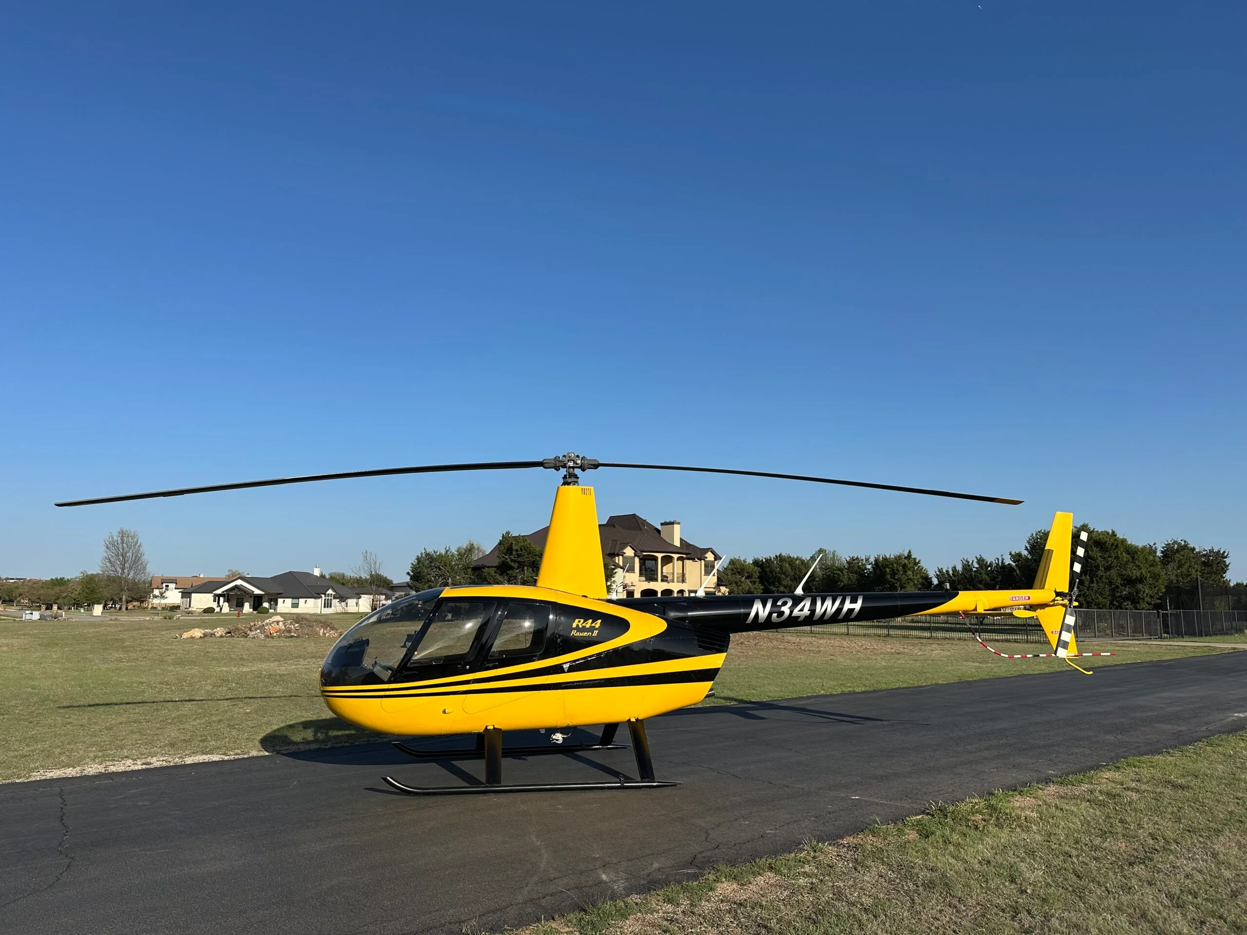 A yellow and black Robinson R44 Raven II helicopter with registration N34WH is parked on an asphalt strip under a clear blue sky.