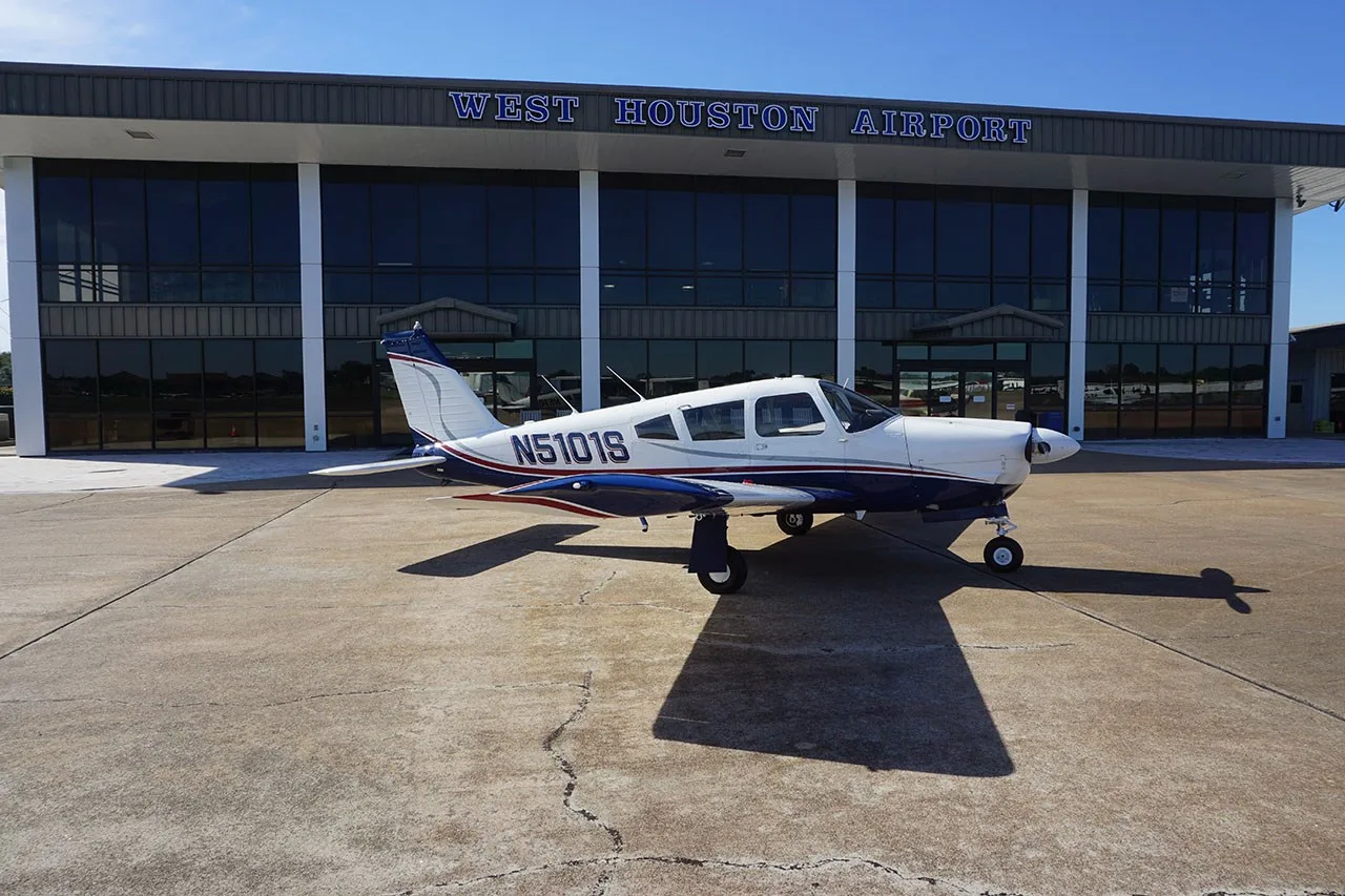 A white and blue Piper PA-28-180 Cherokee Challenger (N5101S) parked on the tarmac in front of the West Houston Airport building.