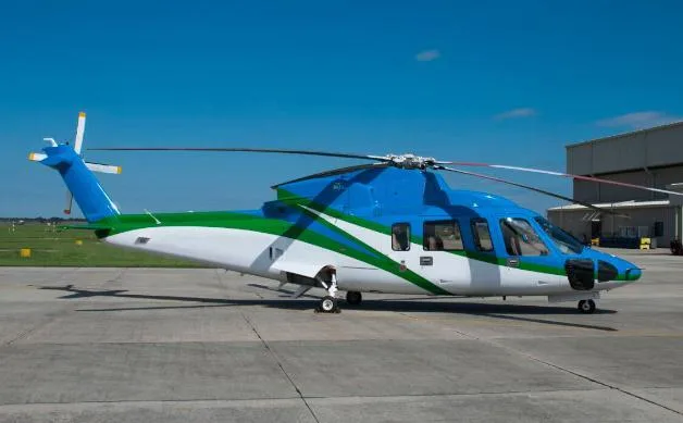 A blue, green, and white AgustaWestland AW139 helicopter parked on a tarmac with a hangar in the background.