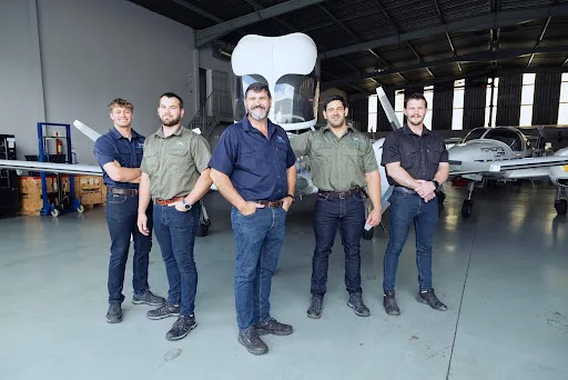 Five men in work shirts and jeans stand in a hangar in front of a white Diamond DA62 aircraft.