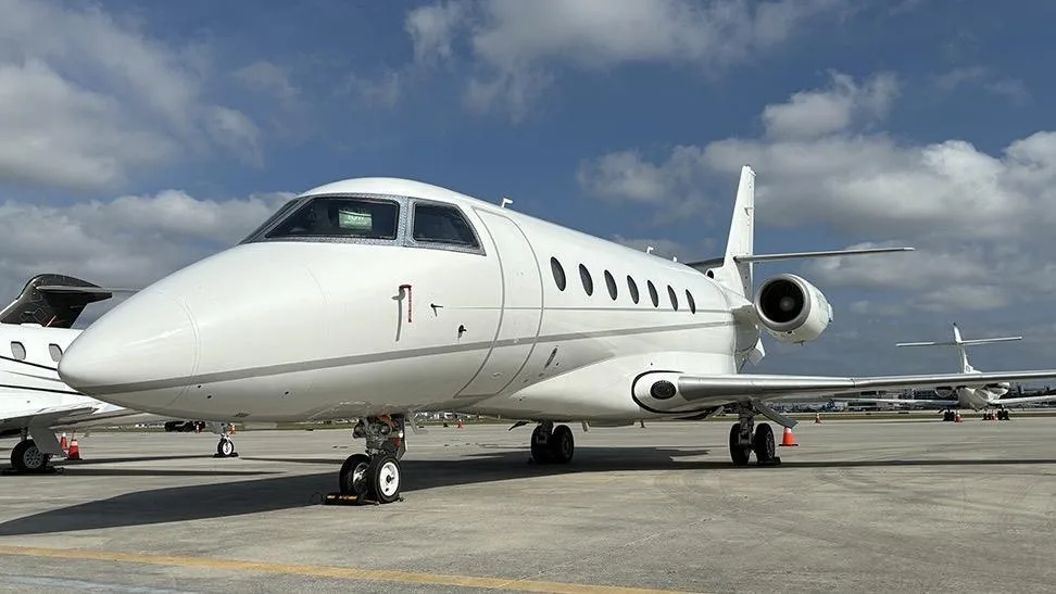A white business jet, likely a Gulfstream, parked on an airport tarmac under a partly cloudy sky.