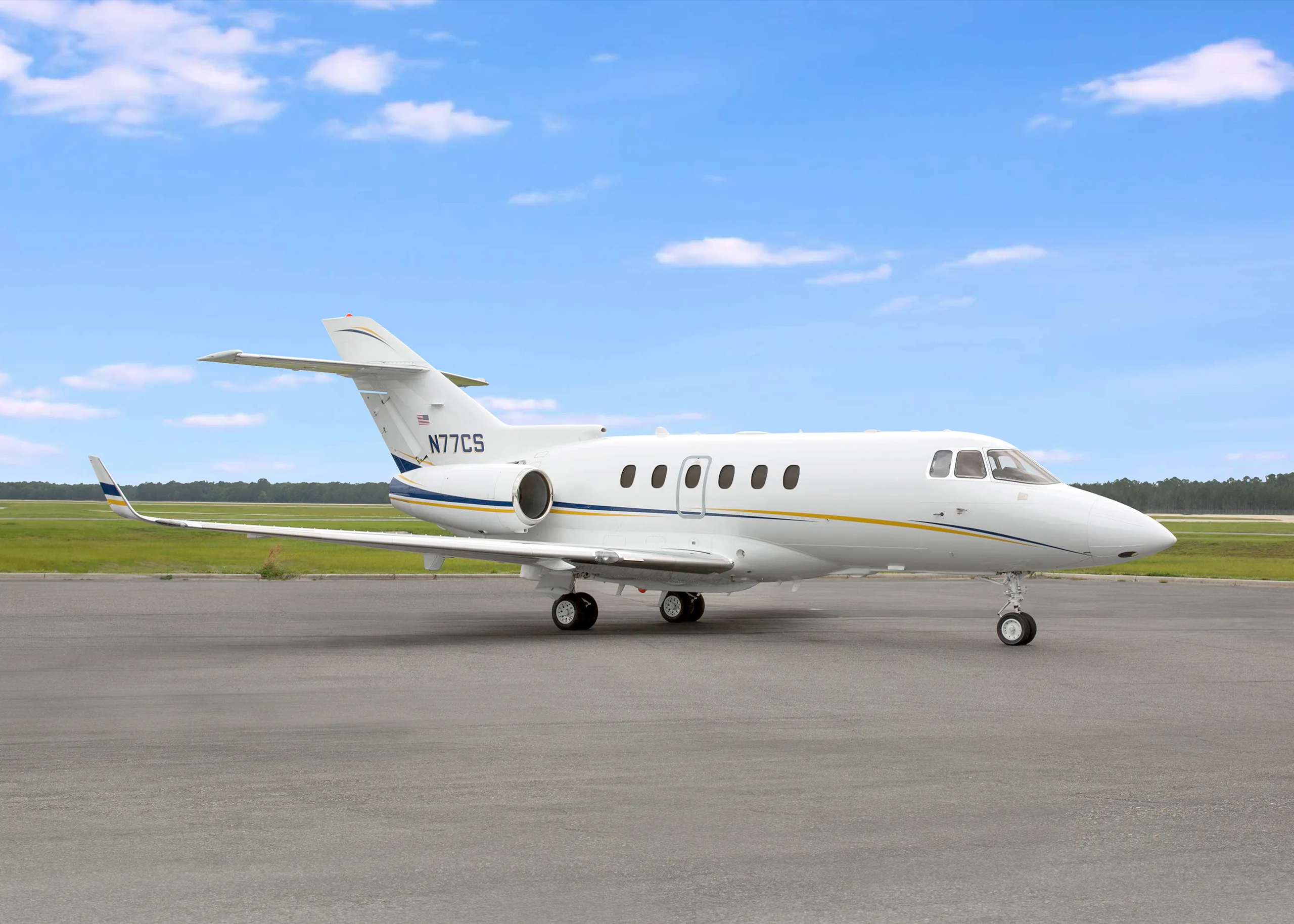 A white business jet with blue and gold stripes, registration N77CS, parked on an asphalt tarmac under a partly cloudy sky.