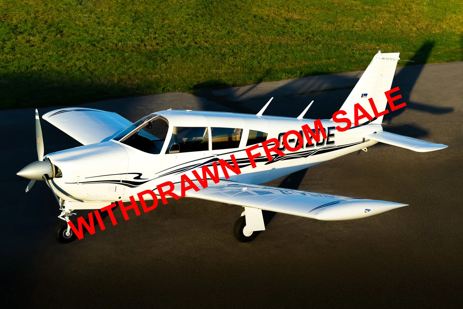 A white single-engine propeller aircraft with black stripe decals is parked on an asphalt runway next to green grass, viewed from above.