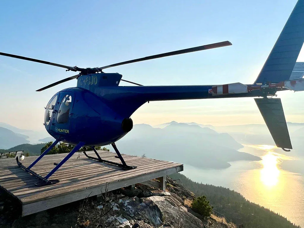 A blue helicopter with registration C-FJJ0 is parked on a wooden helipad on a mountain overlooking a sunlit bay at sunset.
