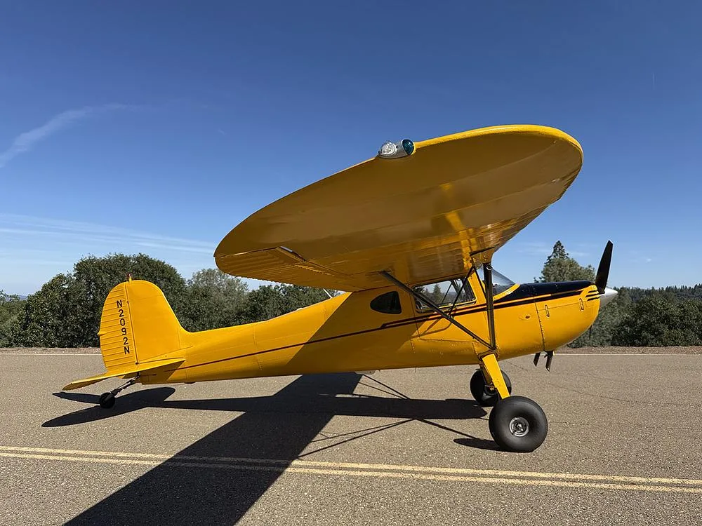 Side view of the yellow high-wing aircraft N2092N, parked on an asphalt surface with a clear blue sky and green trees.