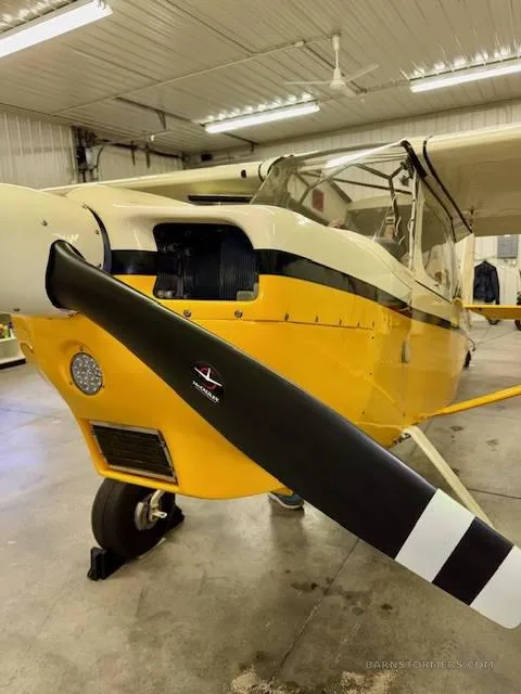 A front-quarter view of a yellow and white high-wing aircraft with a black propeller, parked inside a hangar.
