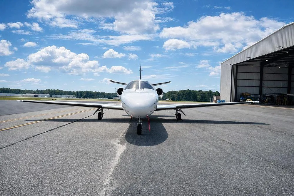 A white business jet, tail number N173AA, viewed head-on on an airfield with a large hangar to the right.