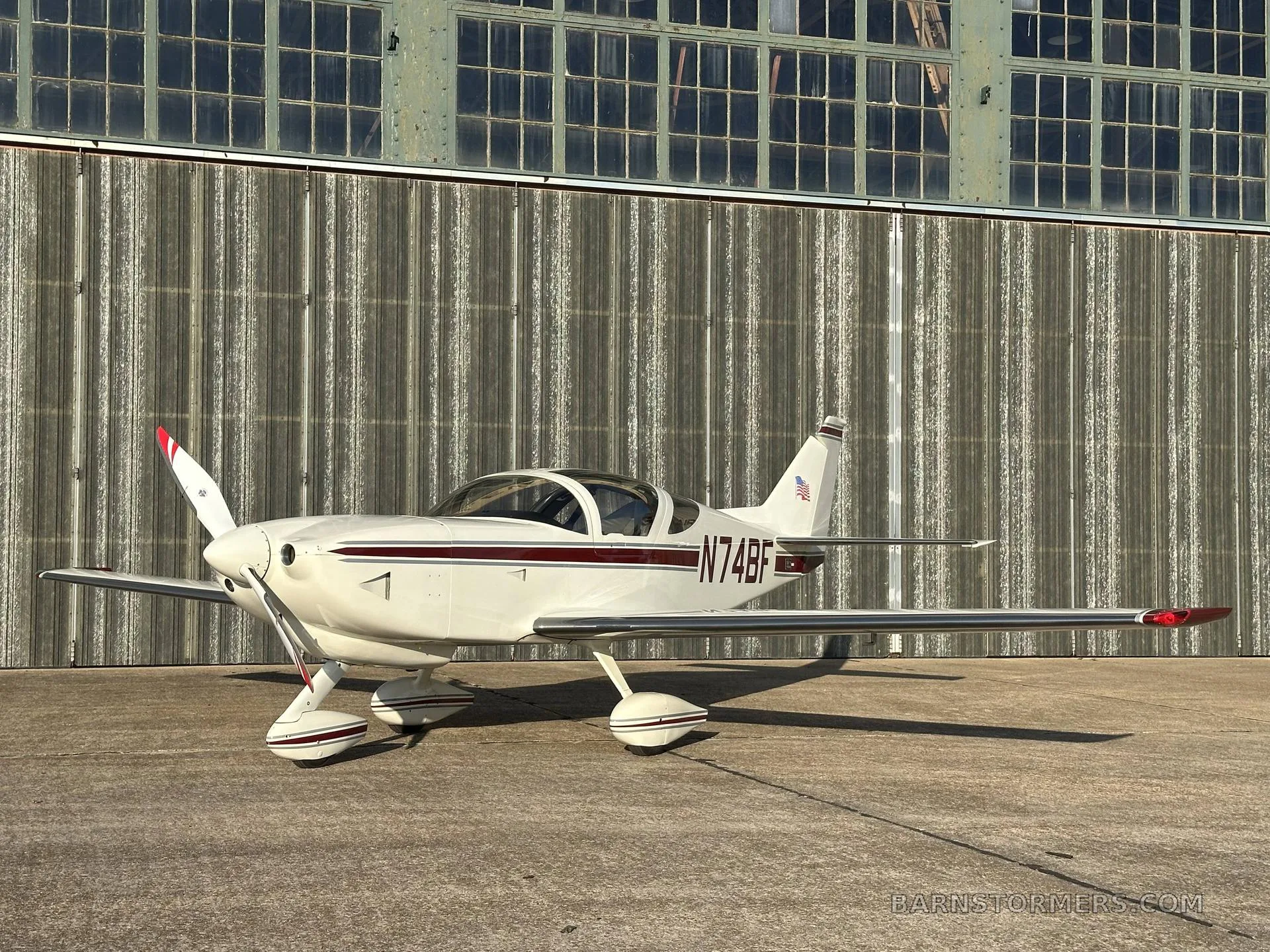 A white single-engine aircraft with a maroon stripe and registration N74BF is parked on concrete in front of a corrugated metal hangar.
