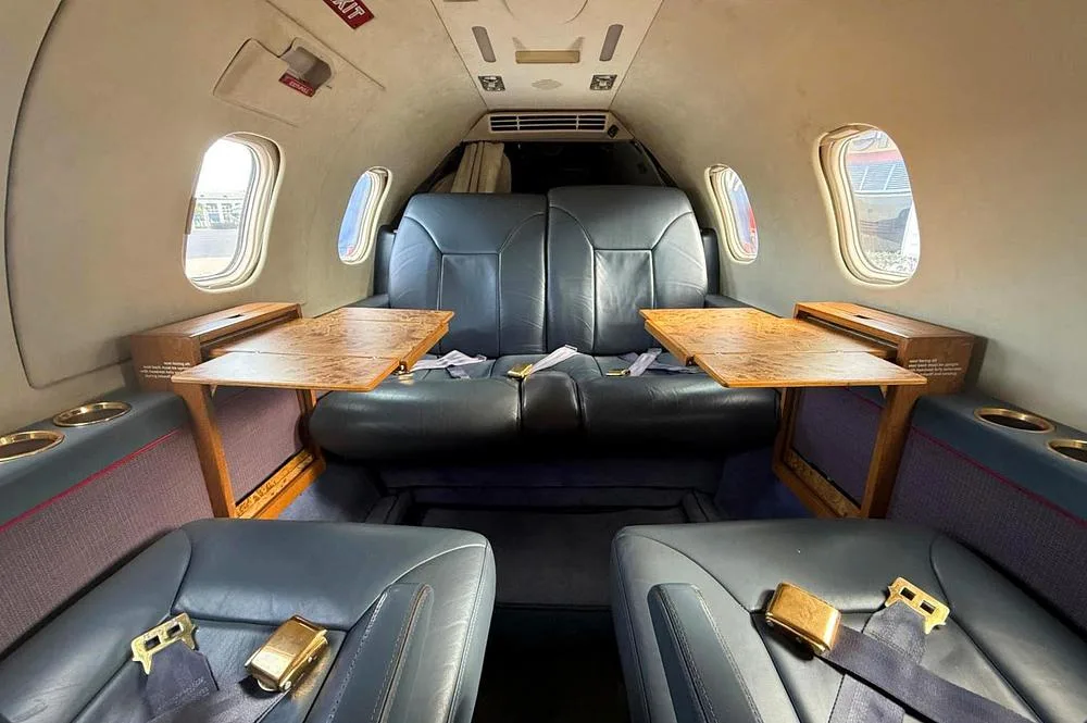 The interior cabin of a private jet featuring dark blue leather seats and wooden fold-out tables.