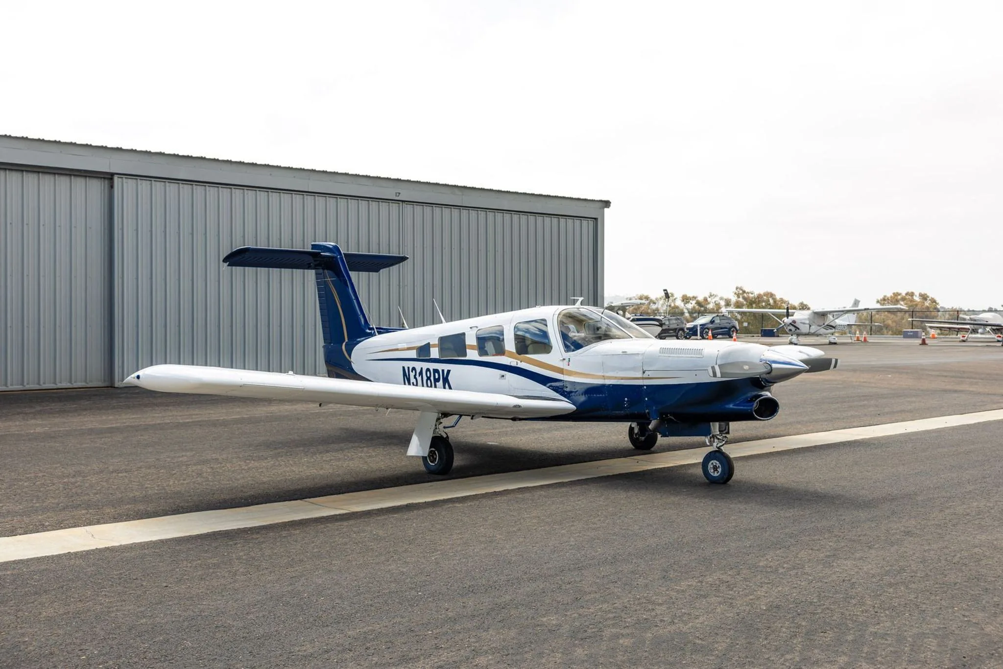 A three-quarter front view of the white and blue Piper aircraft N318PK parked on an airport tarmac with other planes in the distance.