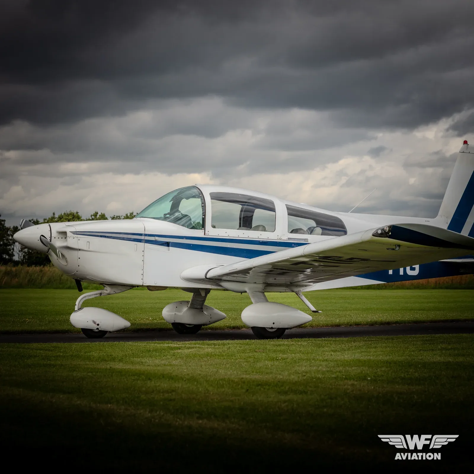 A white single-engine aircraft with blue stripes on its fuselage and tail, parked on a grassy airfield under a cloudy sky.