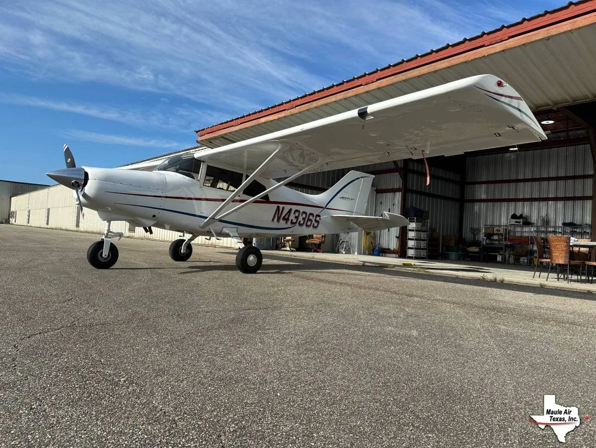 A white single-engine propeller aircraft with red and blue stripes, registration N4336S, parked on an asphalt tarmac next to an open hangar.