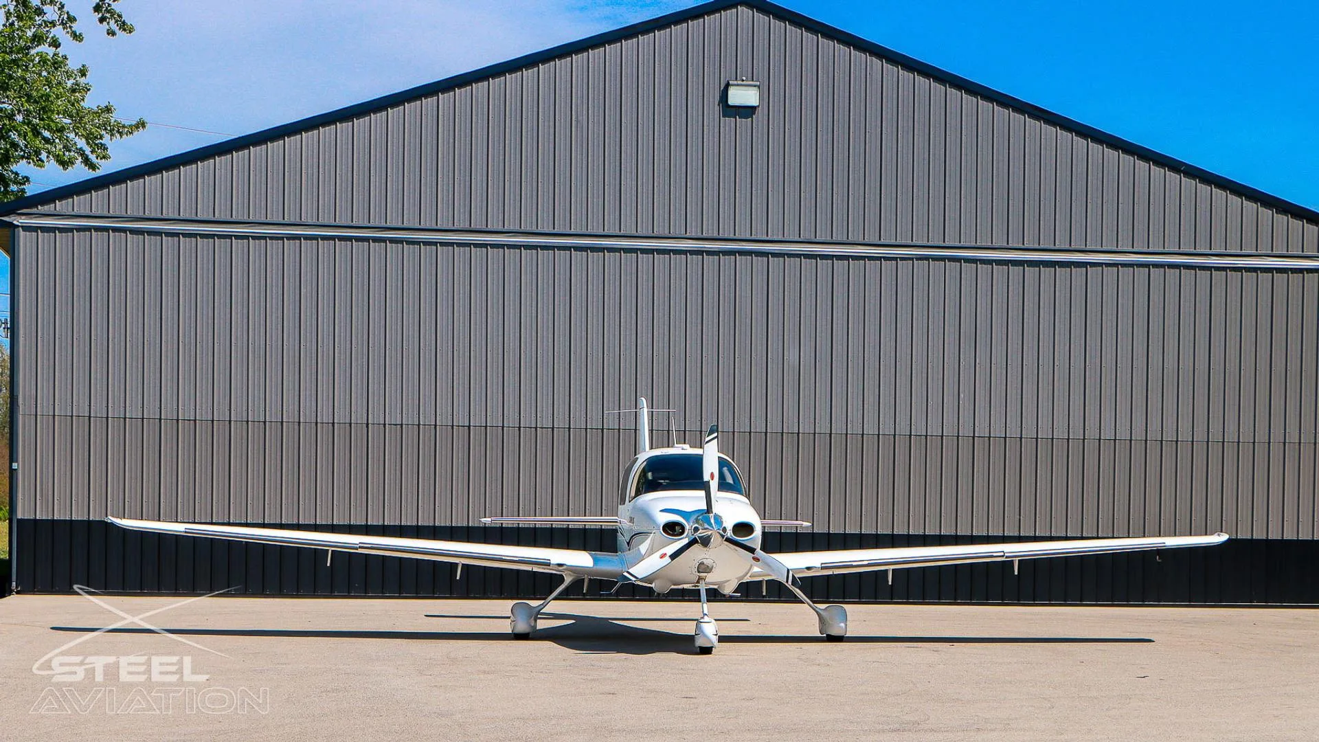 Front view of a white Cirrus SR22 aircraft with black and grey stripes, parked on an tarmac in front of a grey hangar.
