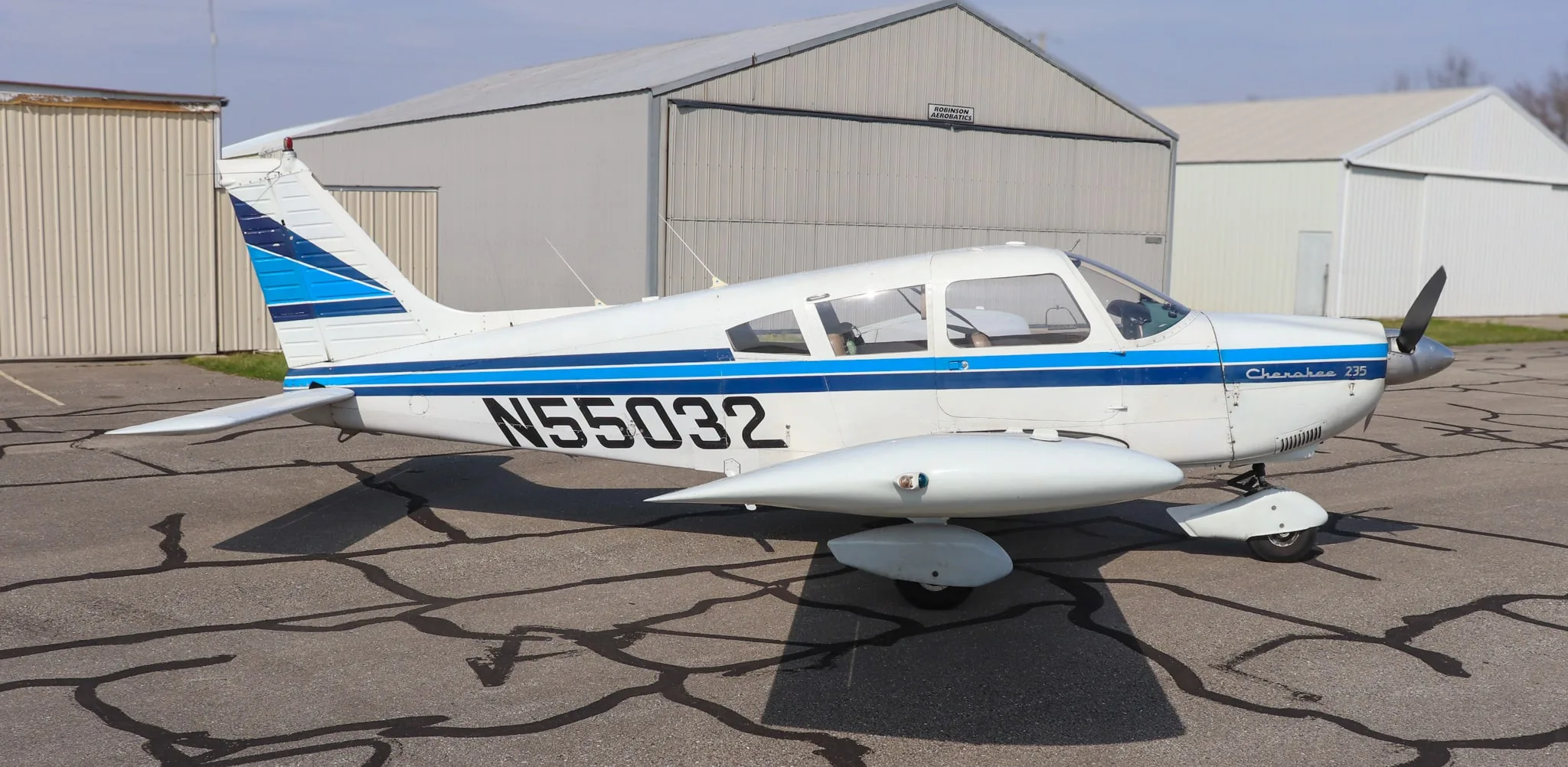 A side view of the white Piper Cherokee 235 aircraft with blue stripes and registration N55032, parked on an asphalt ramp.